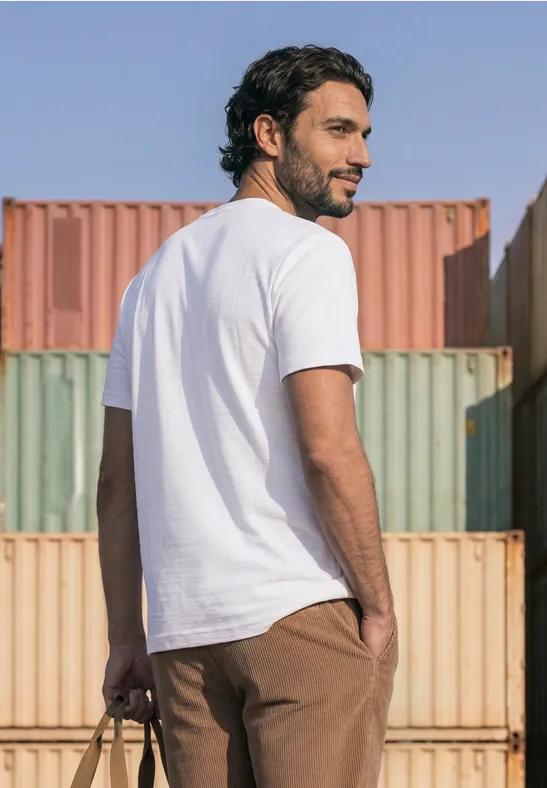 Een man in een wit t-shirt en bruine broek staat voor kleurrijke containers, kijkt over zijn schouder.
