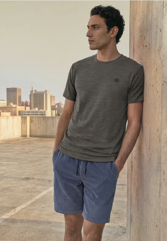 Een man leunt tegen een betonnen pilaar op een dak, draagt t-shirt en korte broek.