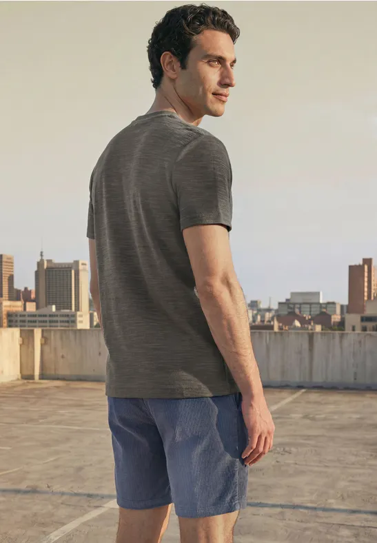 Een man in een grijs T-shirt en blauwe short staat op een dak met stadszicht en kijkt over zijn schouder.