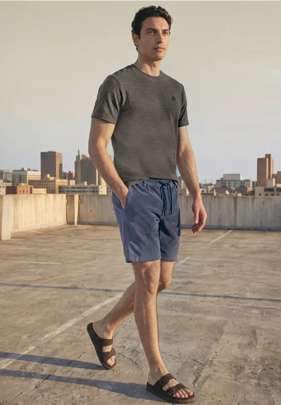 Een man staat op een parkeerdak, draagt een grijs T-shirt, blauwe short en sandalen, met een skyline op de achtergrond.