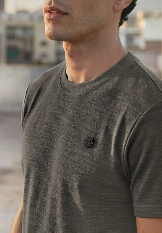 Een man draagt een grijs T-shirt met klein borstlogo, vage stedelijke achtergrond.