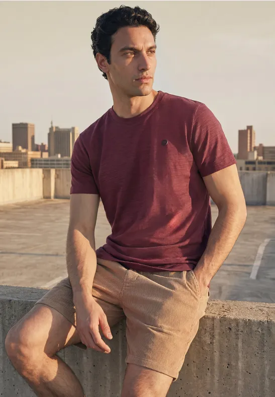 Een man zit op de rand van een dak, draagt rood T-shirt en beige korte broek, starend in de verte.