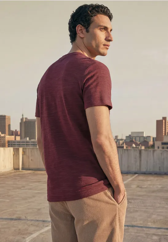 Een man staat op een dak, kijkt over de stad en draagt een roodbruin T-shirt met beige broek.