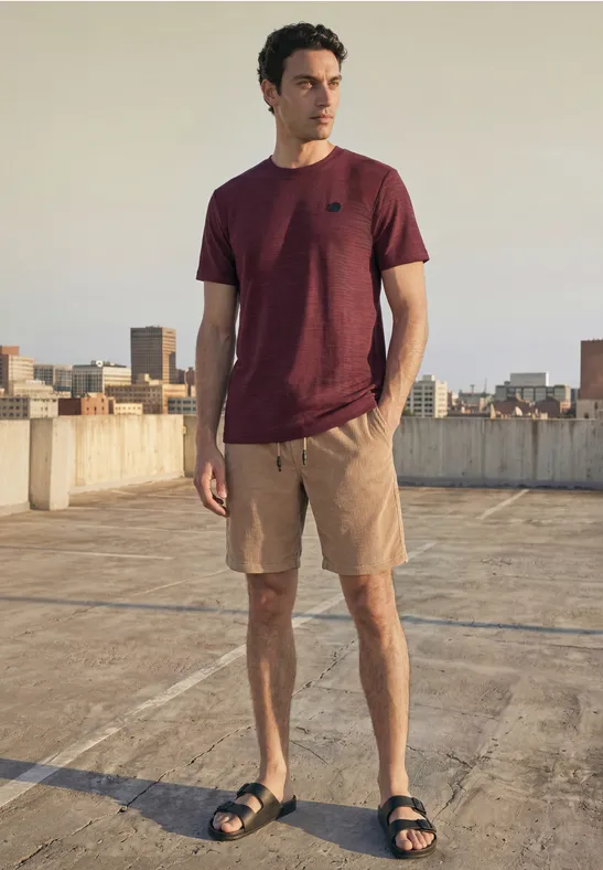 Een man op het dak van een parkeergarage met stadszicht draagt een bordeauxrood T-shirt, korte broek en sandalen.