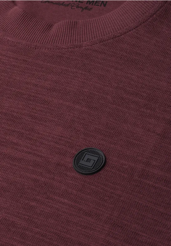Bordeauxrood gebreid T-shirt met zwart rond logo-embleem op de borst.