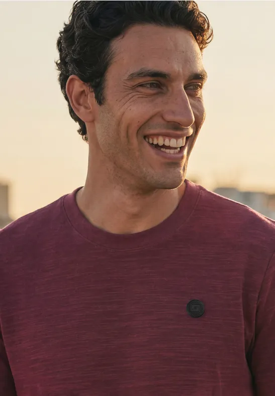 Een lachende man draagt een bordeauxrood t-shirt met klein logo.