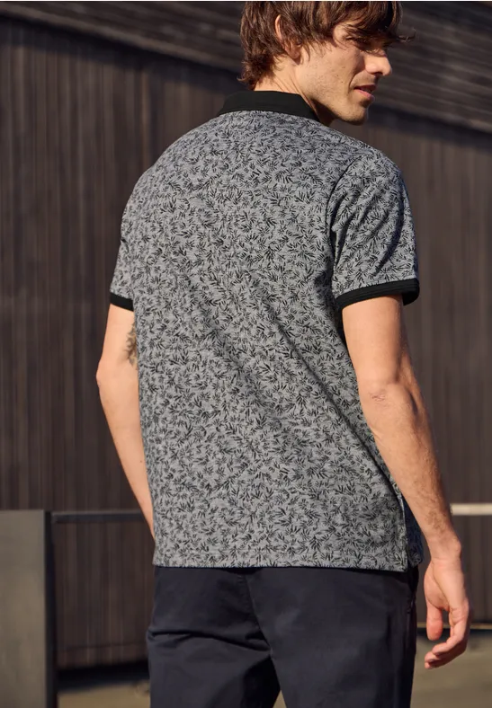 Ein Mann steht von hinten vor einer Holzverkleidung, trägt ein gemustertes T-Shirt und dunkle Hose.