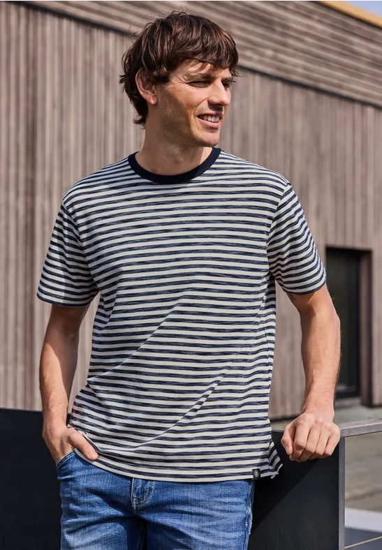 Junger Mann in gestreiftem T-Shirt und Jeans lehnt an Holzgeländer vor modernem Gebäude.