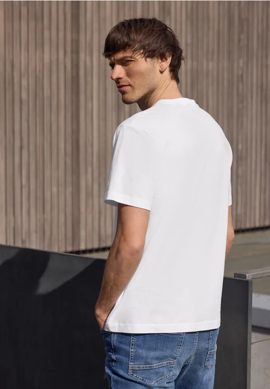 Ein junger Mann steht seitlich vor Holzwand, trägt weißes T-Shirt und blaue Jeans.