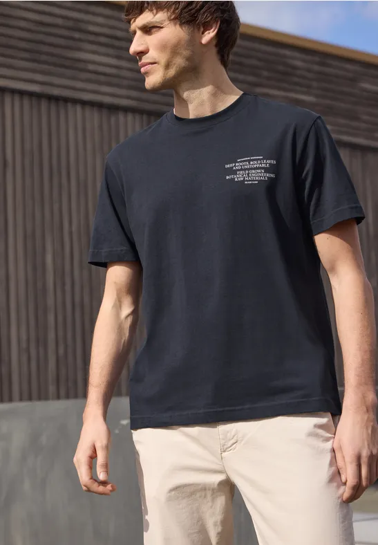 Ein junger Mann mit schwarzem T-Shirt und hellen Hosen steht vor einem Holzgebäude im Freien.