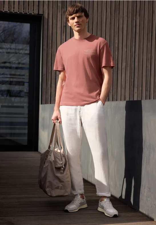 Mann lehnt an Holzfassade, trägt rotes T-Shirt, helle Leinenhose, weiße Sneaker und hält eine Tasche.