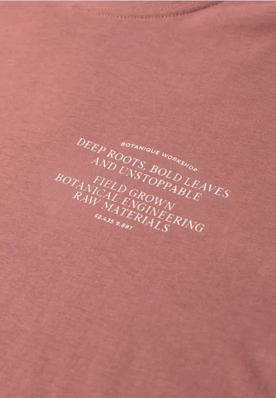 Rosa T-Shirt mit weißem Slogan über tiefe Wurzeln, starke Blätter und nachhaltige Materialien.