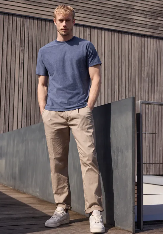 Ein Mann lehnt an einem Betongeländer vor einer Holzfassade, trägt blaues T-Shirt, helle Chinohose und Sneaker.