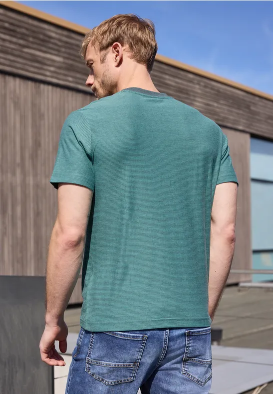 Mann mit grünem T-Shirt und blauer Jeans steht rückwärts vor Holzfassade unter blauem Himmel.