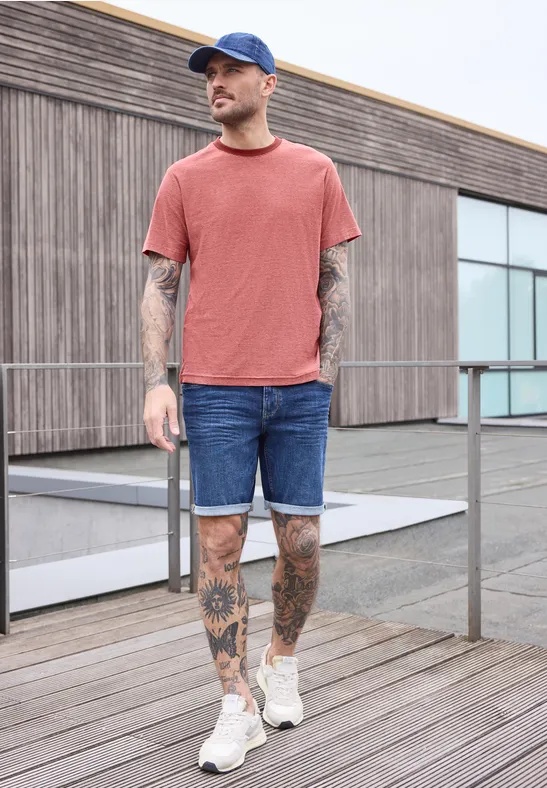 Ein Mann mit Tattoos trägt rotes T-Shirt, Jeans-Shorts und weiße Sneaker auf einer Holzterrasse.