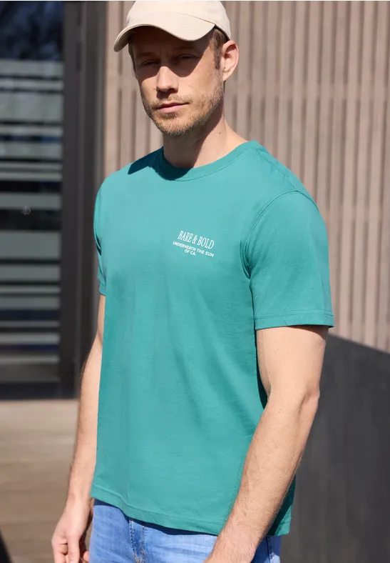 Ein junger Mann trägt draußen ein türkisfarbenes T-Shirt und eine Jeans.