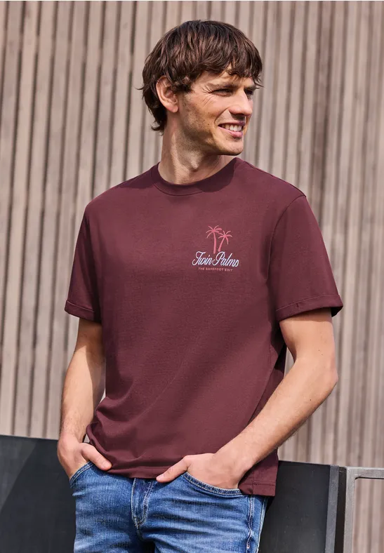 Un homme en t-shirt bordeaux et jean, debout devant une façade en bois, regarde sur le côté.