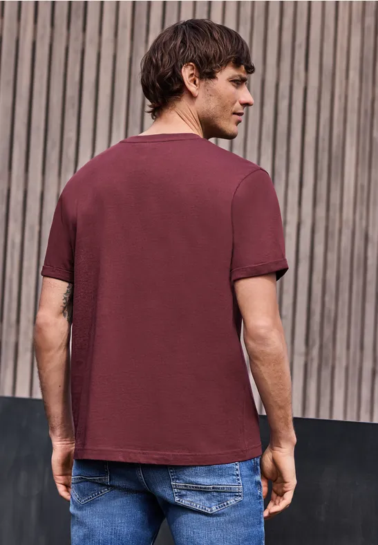 Un homme vu de dos en t-shirt bordeaux et jean, se tient devant un mur de lattes en bois.