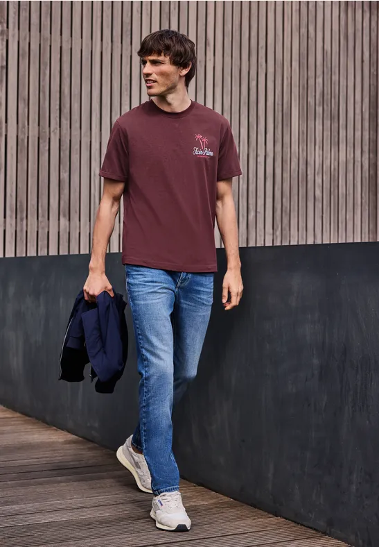 Un homme en jean et t-shirt bordeaux marche devant une façade en bois en tenant une veste.