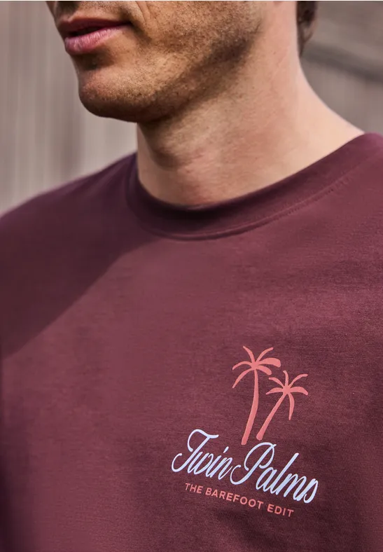 Gros plan d’un homme portant un t-shirt bordeaux avec motif de palmiers et inscription «Twin Palms»