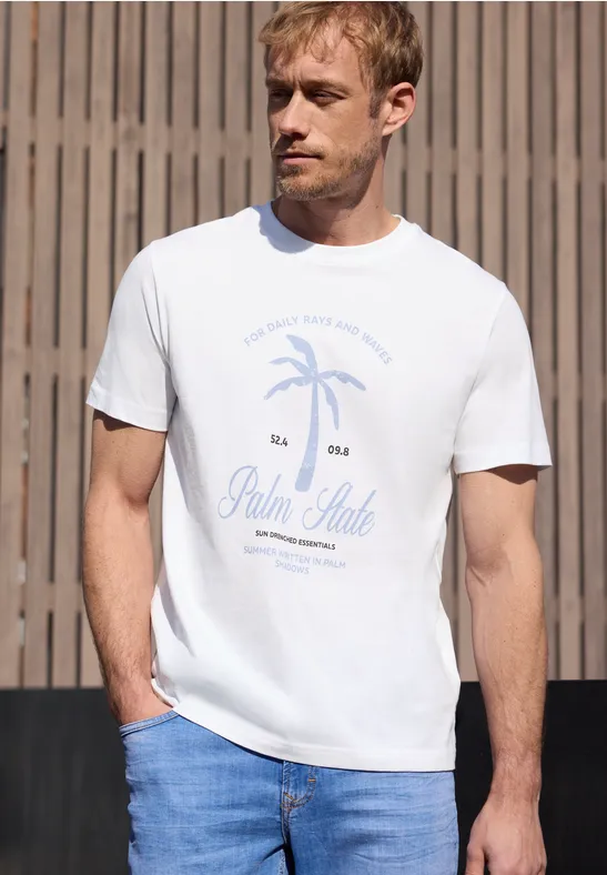 Ein Mann trägt ein weißes T-Shirt mit Palmenmotiv und Schriftzug 'Palm State' sowie Jeans vor Holzwand.