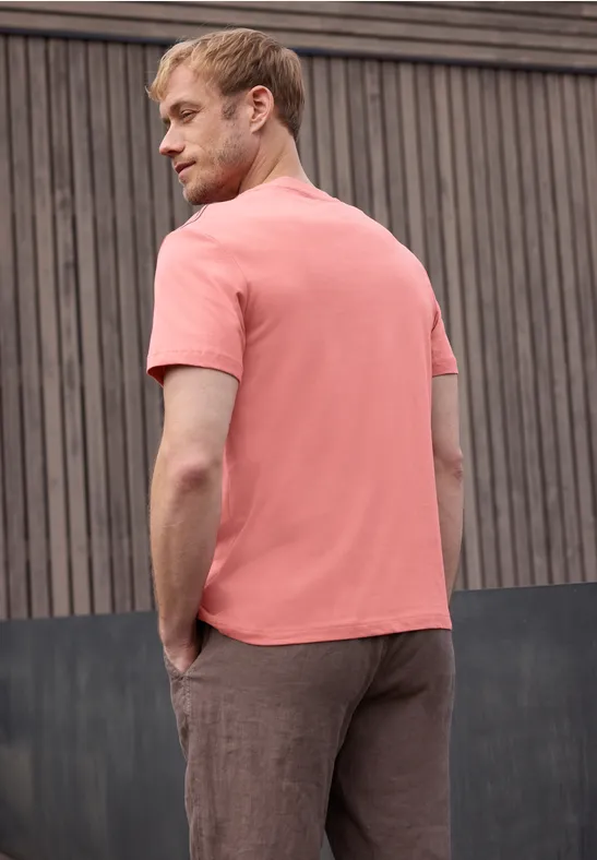 Mann in rostrotem T-Shirt und brauner Hose lehnt seitlich an einer Holzverkleidung.