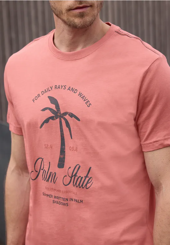 Ein Mann trägt ein lachsfarbenes T-Shirt mit einem Palmenmotiv und Schriftzug „Palm State“.