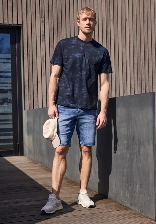 Ein Mann steht auf einer Holzterrasse, trägt dunkles T-Shirt, Jeans-Shorts, weiße Sneaker und hält eine beige Kappe.