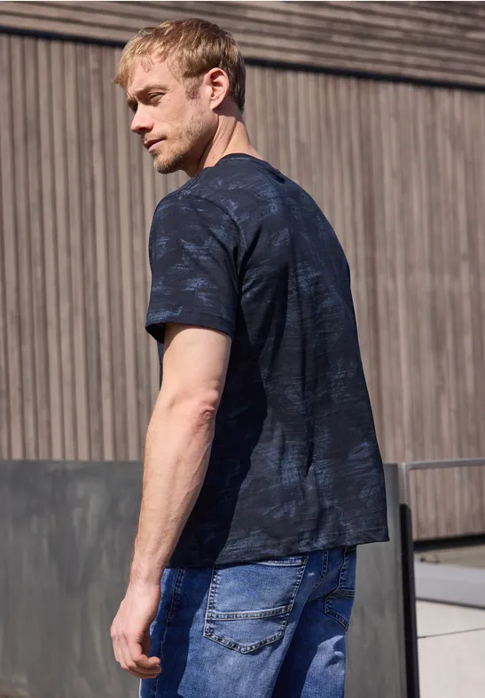 Ein Mann mit schwarzem T-Shirt und blauen Jeans steht vor einer Holzfassade.