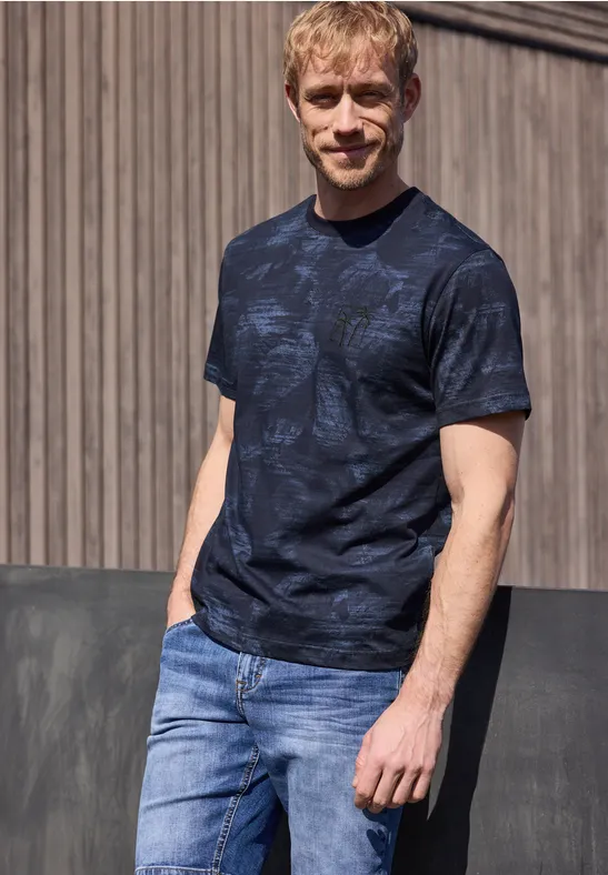 Ein Mann trägt ein dunkelblaues T-Shirt mit Muster und hellblaue Jeans, lehnt an einer Holzvertäfelung.