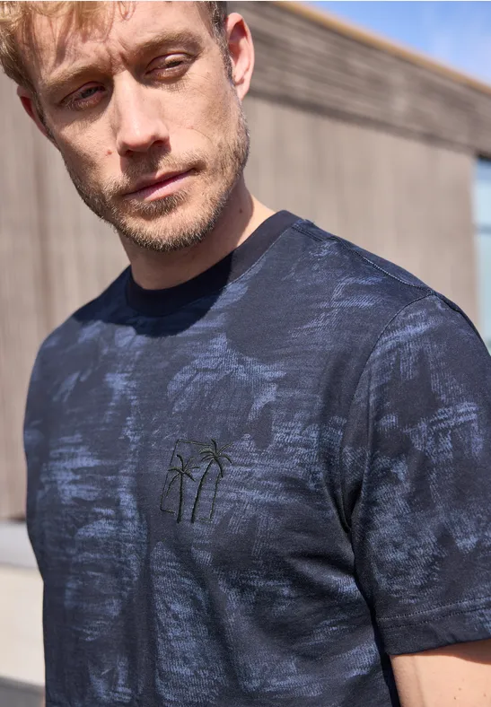 Mann trägt dunkelblaues T-Shirt mit Palmenstickerei, stehend vor einer Holzwand im Freien.
