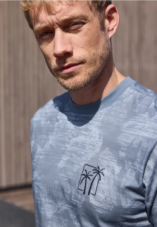 Ein blonder Mann trägt ein blaues T-Shirt mit Palmen-Logo, stehend vor einer Holzverkleidung.