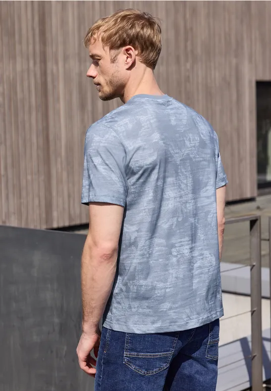 Ein Mann von hinten in einem grau-blauen T-Shirt steht vor einer Holzwand im Freien.