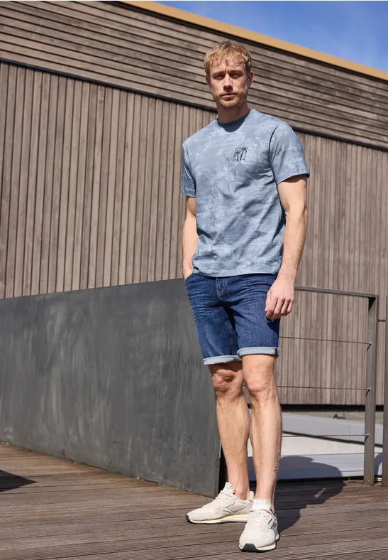 Ein Mann trägt ein gemustertes T-Shirt, Jeans-Shorts und weiße Sneakers auf einer Holzterrasse.