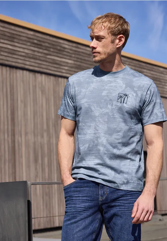 Ein Mann steht vor einer Holzwand, trägt ein graues Batik-T-Shirt und Jeans, Hände in den Taschen.