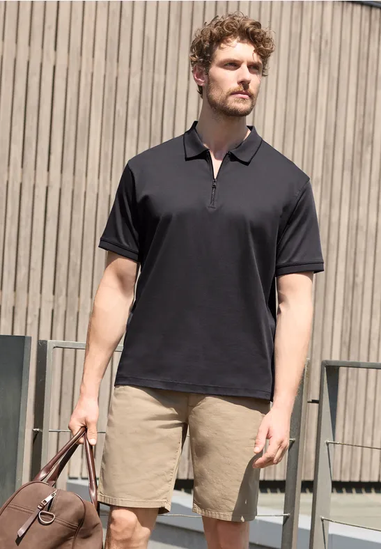 Ein Mann mit schwarzem Poloshirt und beigefarbenen Shorts hält eine Tasche vor einer Holzfassade.