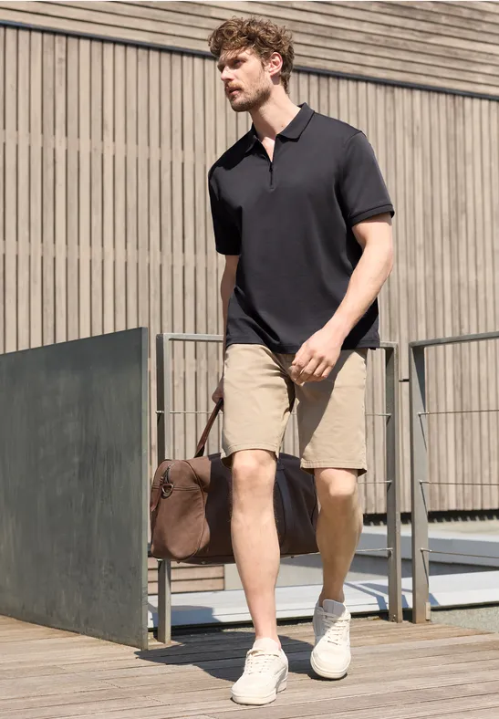 Ein Mann in schwarzem Polo, khakifarbenen Shorts und weißen Sneakers trägt eine Reisetasche und geht an einem modernen Gebäude vorbei.
