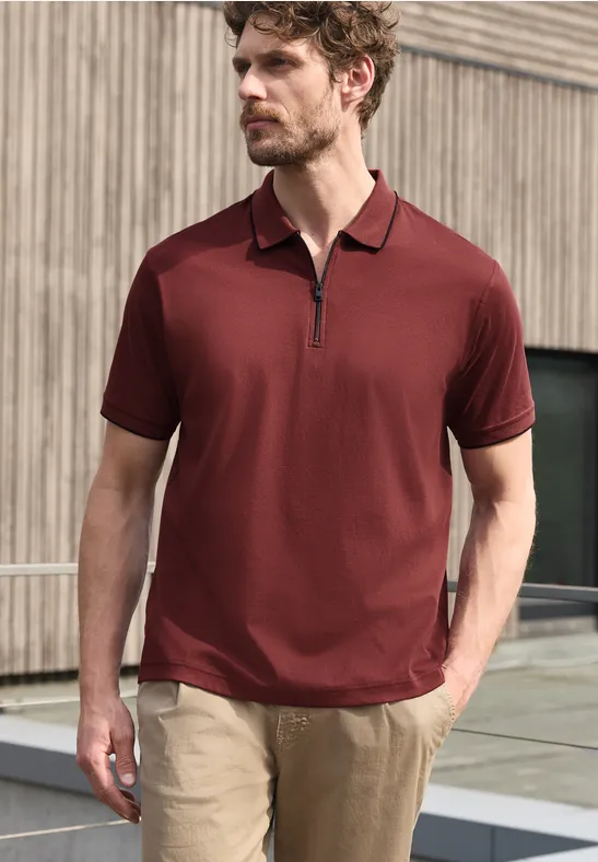 Ein Mann trägt ein bordeauxrotes Kurzarm-Poloshirt mit Reißverschluss und helle Hose vor einer Holzfassade.