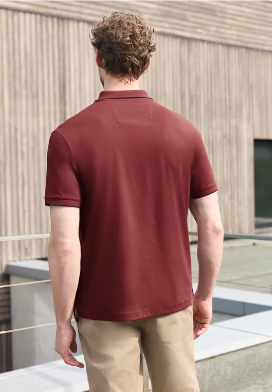 Ein Mann von hinten in einem weinroten Poloshirt und hellen Hosen steht vor einer Holzfassade.