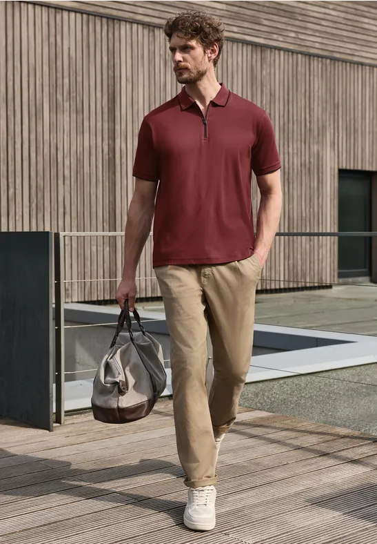 Mann mit rotem Poloshirt, beigen Hosen und weißen Sneakers trägt Reisetasche und geht an moderner Fassade vorbei.