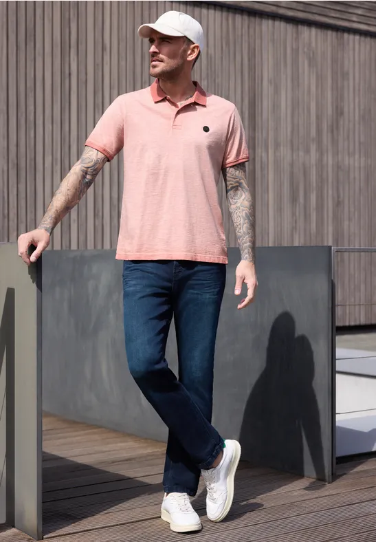 Ein Mann mit Tattoos trägt rosa Polo, Jeans und weiße Sneaker, lehnt lässig an einer Holzfassade.