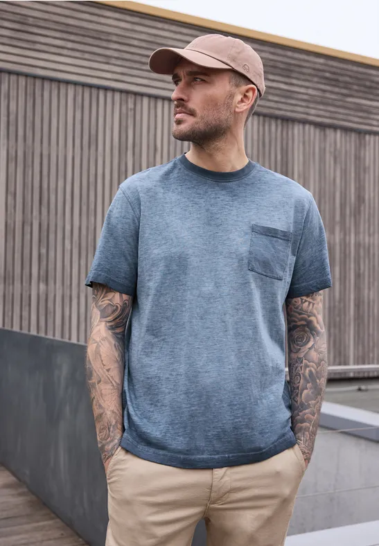 Ein Mann mit Tattoo-ärmeln in blauem T-Shirt und Kappe steht vor einer Holzwand.