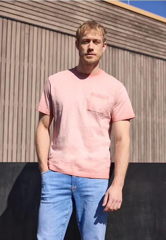 Ein Mann in rosa T-Shirt und blauen Jeans steht vor einer Holzfassade.