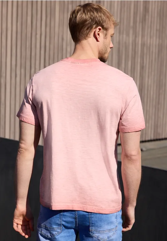 Ein Mann steht im Freien, Rücken zu sehen, trägt ein rosa T-Shirt und blaue Jeans vor einer Holzpaneelwand.
