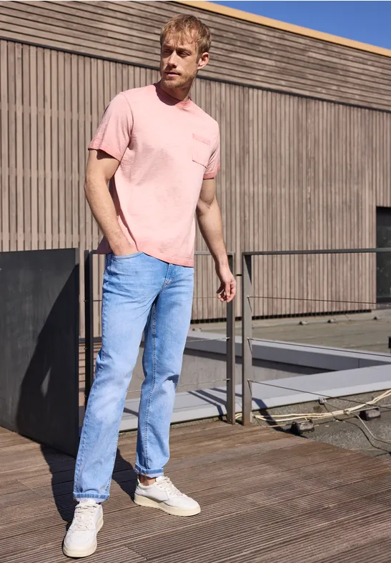 Ein Mann in rosa T-Shirt, hellblauen Jeans und weißen Sneakern steht auf einer Holzterrasse vor einem Haus.