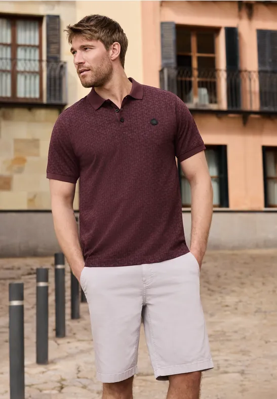 Ein Mann trägt ein dunkelrotes Polohemd und graue Shorts vor einem Altbau.