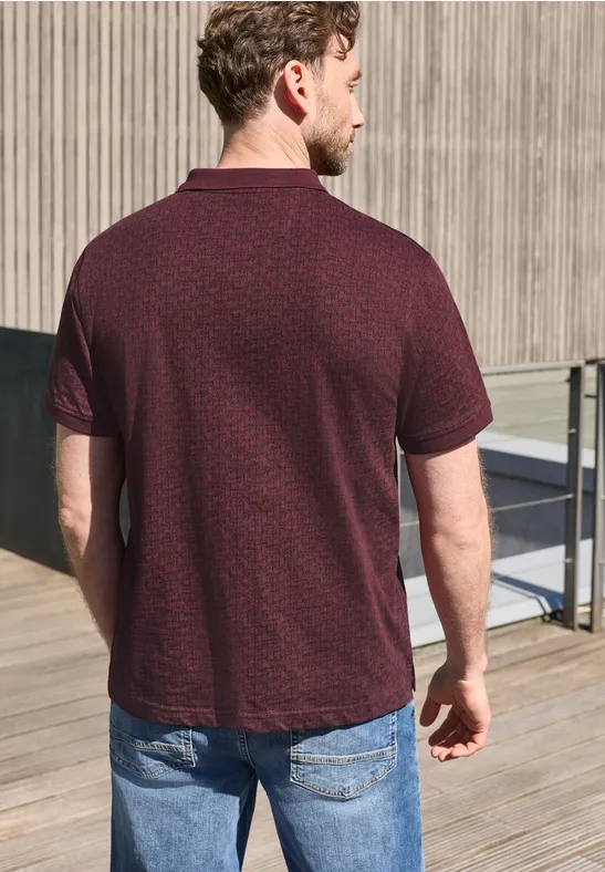 Ein Mann von hinten in burgunderfarbenem Poloshirt mit dezenter Musterung und Jeans auf einer Holzterrasse.