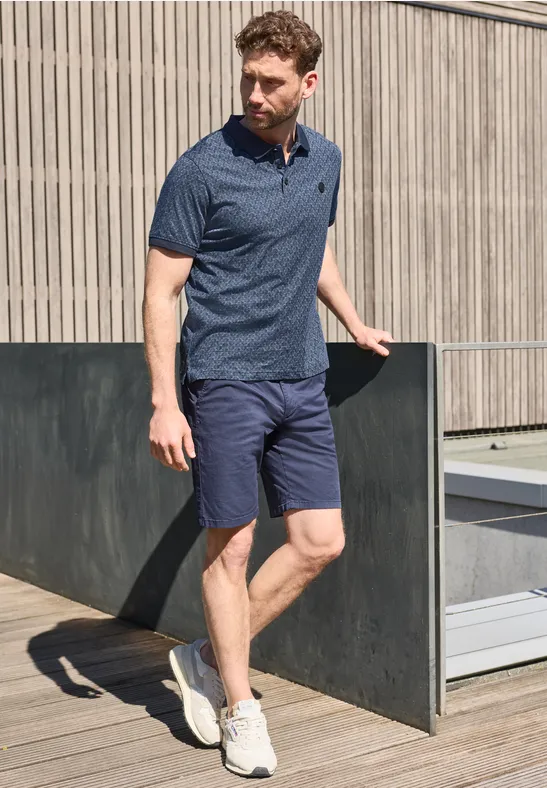 Een man leunt op een dakterras leuning, draagt blauw poloshirt, korte broek en witte sneakers.