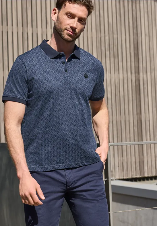 Een man staat voor een houten gevel en draagt een blauw poloshirt met motief en een donkerblauwe broek.