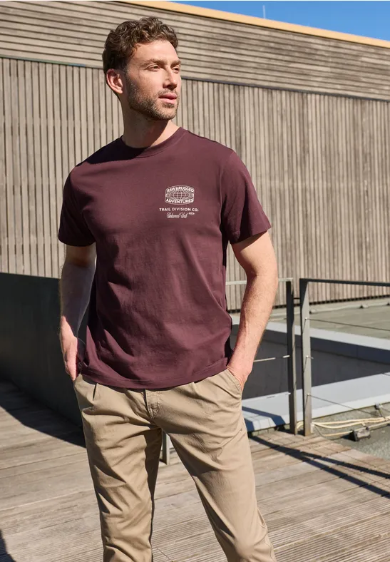 Een jonge man staat op een dak voor een houten gevel, gekleed in een t-shirt en chino’s.